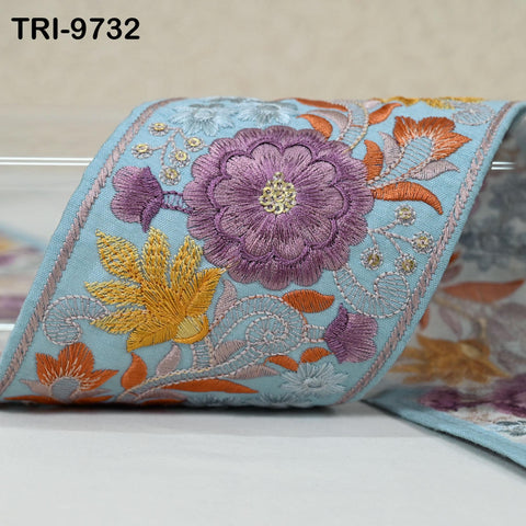 a close-up view of a decorative fabric with a floral pattern. The fabric is adorned with a variety of colorful flowers, including purple, orange, and yellow flowers, as well as leaves and other decorative elements. The fabric appears to be a piece of embroidered or sewn fabric, showcasing the intricate and vibrant design.
