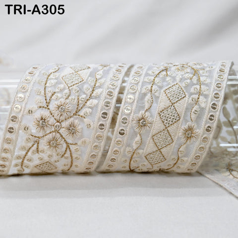 a close-up view of a decorative white fabric with intricate gold embroidery patterns. The fabric appears to be a ribbon or lace, with a repeating floral and diamond-shaped motif. The image does not contain any text or other objects, and the focus is solely on the detailed embroidery work.