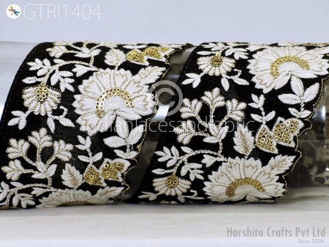a close-up view of a decorative fabric with a floral pattern in black and white, accented with gold elements. The fabric appears to be a piece of lace or a similar type of textile, with intricate embroidered designs. The background is a plain white surface, which allows the focus to remain on the fabric and its details.