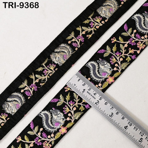 a decorative fabric with a floral pattern, including a black background with a repeating design of purple and green flowers and a gray squirrel. A ruler is placed on the fabric, indicating the size of the design.