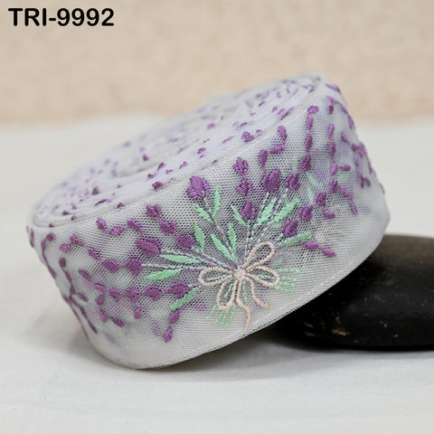 a close-up view of a decorative fabric with a floral pattern, featuring purple and green elements. The fabric appears to be a piece of lace or a similar type of material, with a floral motif and a ribbon-like texture. The fabric is placed on a surface, possibly a table or a countertop, and there is a small black object, possibly a stone or a pebble, located next to it.