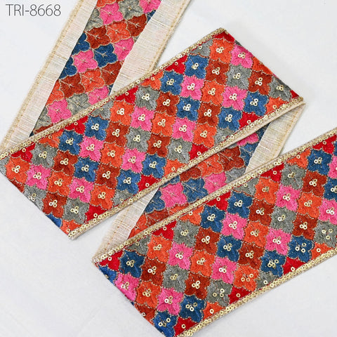 a decorative fabric with a vibrant, colorful pattern of squares and rectangles, adorned with sequins and embellishments. The fabric appears to be a decorative trim or border, with a repeating pattern of red, blue, and pink squares and rectangles. The fabric is displayed against a plain white background, highlighting its intricate design and vibrant colors.
