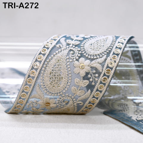 a decorative fabric with intricate embroidered patterns in shades of blue and white. The fabric appears to be a decorative ribbon or trim, with a floral and paisley motif. The background is a plain white surface, which highlights the vibrant colors and patterns of the fabric.
