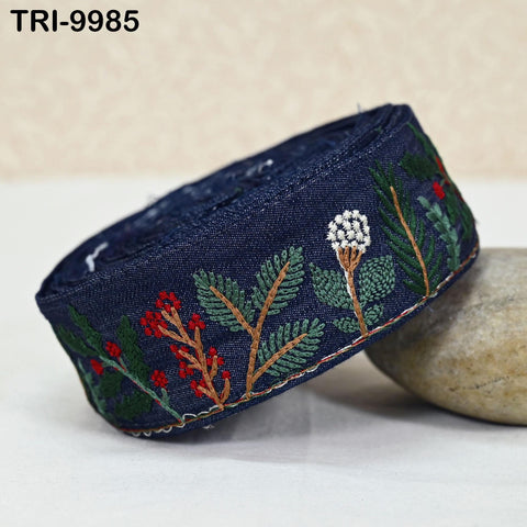 A close-up image of a blue fabric with a floral embroidered design, including red berries and green leaves, resting on a white surface.