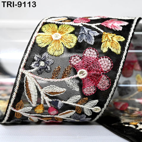a close-up view of a decorative fabric with a floral pattern. The fabric has a black background and is adorned with a variety of colorful flowers and leaves, including yellow, pink, and purple flowers. The fabric appears to be a decorative piece, possibly used for embellishment or as a textile material.