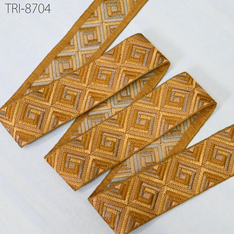 a piece of fabric with a geometric pattern in shades of gold and brown, featuring a repeating diamond-shaped motif. The fabric appears to be a decorative textile, possibly used for embellishment or clothing.