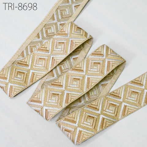a close-up view of a decorative fabric with a geometric pattern in shades of gold and beige. The fabric appears to be a decorative trim or ribbon, with a repeating diamond pattern. The fabric is neatly arranged on a white surface, creating a visually appealing contrast.