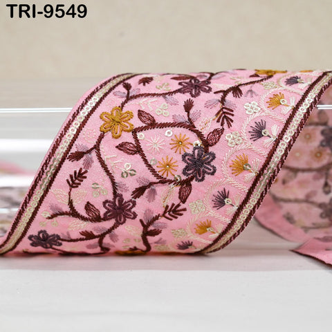 a close-up view of a decorative fabric with a floral pattern in shades of pink and purple. The fabric appears to be a decorative ribbon or textile, with intricate embroidered designs and a metallic trim.