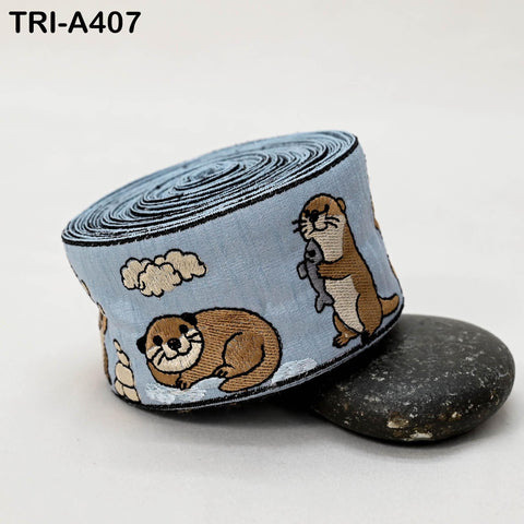 A blue and white embroidered fabric with a playful design of cartoon otters, including one holding a fish. The fabric is rolled up and placed on a black rock.