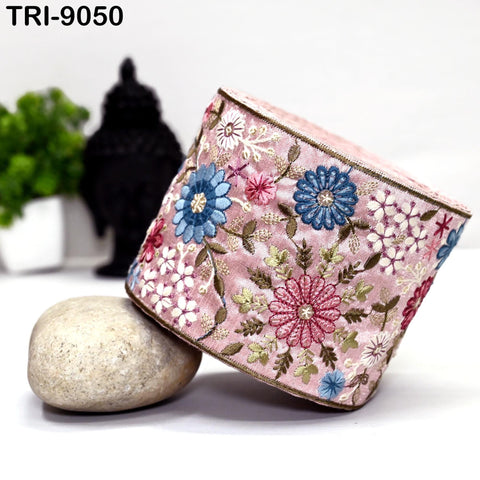 a pink and blue floral embroidered object, which appears to be a decorative piece, placed on a white surface. In the background, there is a black object and a white object, possibly a plant or a decorative item.