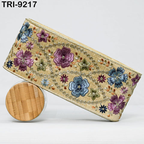 a decorative fabric with a floral pattern, including various shades of purple, blue, and green flowers, as well as some brown and beige elements. The fabric appears to be a decorative trim or border, possibly used for embellishing a piece of clothing or accessory.