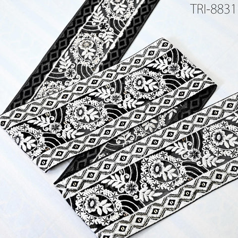 a close-up view of a decorative fabric with a black and white floral pattern. The fabric appears to be a decorative trim or ribbon, with a repeating pattern of floral and geometric designs. The fabric is placed on a white surface, which highlights its intricate details and colors.