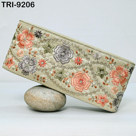 a decorative beige fabric with a floral pattern, including various shades of orange, pink, and green flowers, as well as smaller floral elements. The fabric is displayed on a flat surface, with a small rock placed underneath it.
