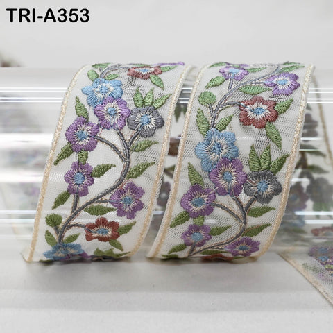 two rolls of decorative fabric with intricate floral embroidery. The fabric has a white background and is adorned with colorful flowers and leaves in shades of purple, blue, and green. The rolls are positioned next to each other, creating a visually appealing display.