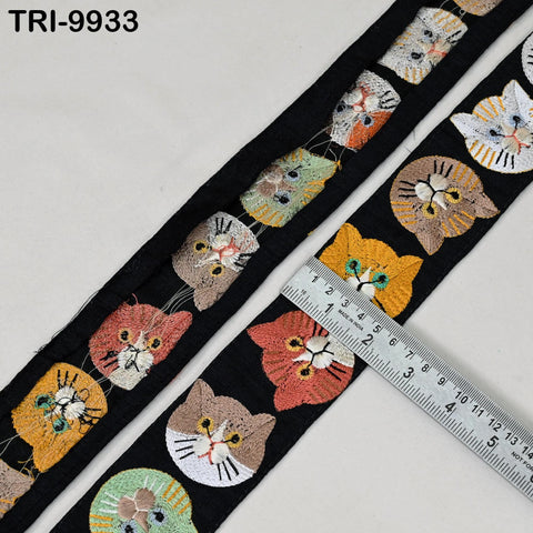 a close-up view of a black fabric with a repeating pattern of colorful cat faces, including various shades of orange, yellow, and gray. A ruler is placed on the fabric, likely for measuring the length or width of the pattern.