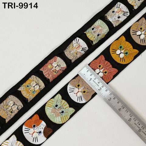 a black ribbon with a repeating pattern of cat faces in various colors, including orange, green, and white. A ruler is placed next to the ribbon to measure its length.