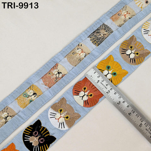 a blue fabric with a repeating pattern of cat faces in various colors, including black, orange, and yellow. A ruler is placed on the fabric, indicating the size of the pattern.