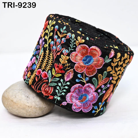 a black fabric with a colorful floral embroidered design, including red, pink, blue, and yellow flowers, as well as green leaves and stems. The fabric is rolled up and placed on a white surface, with a small rock nearby.