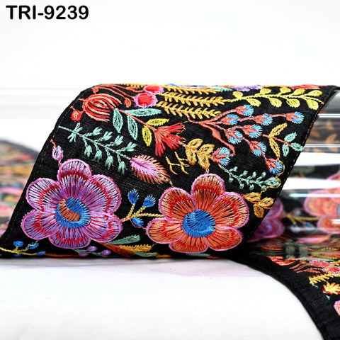 a close-up view of a decorative fabric with a floral pattern. The fabric appears to be black and is adorned with colorful flowers and leaves in shades of red, orange, and yellow. The fabric is folded and placed on a white surface, creating a visually striking contrast between the vibrant colors and the neutral background.