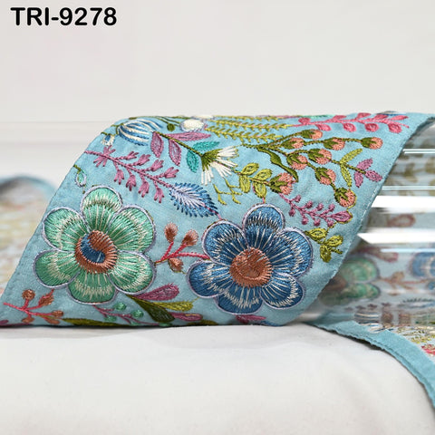 a close-up view of a decorative fabric with a floral pattern, including blue, green, and pink flowers and leaves. The fabric appears to be a piece of embroidered material, possibly used for a decorative purpose or as a textile product.