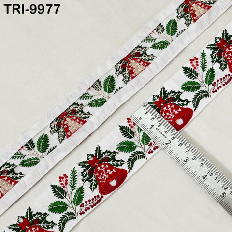a close-up view of a decorative ribbon with a repeating pattern of red and green leaves, along with red bell-shaped embellishments. The ribbon is laid out on a white surface, and a ruler is placed next to it, indicating its size.