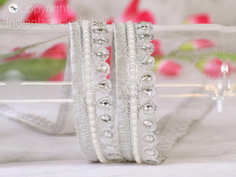 A decorative silver and white lace trim with small rhinestones and pearls is displayed on a white surface, with a blurred background of pink flowers.