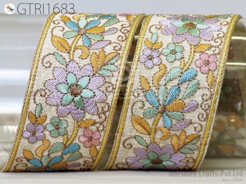 two decorative fabric pieces with intricate floral embroidery in shades of purple, green, and yellow. The pieces are arranged diagonally, with the left piece positioned slightly higher than the right one. The background appears to be a plain white surface, which allows the colorful embroidery to stand out prominently.