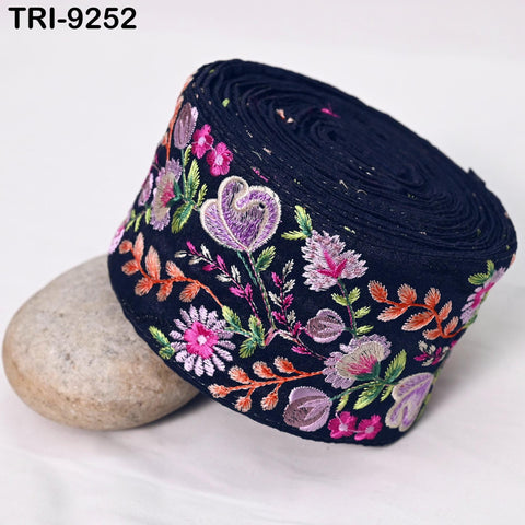 a close-up view of a black fabric with a floral embroidered design, including purple and pink flowers, along with green leaves and branches. The fabric appears to be rolled up and placed on a white surface, with a rock visible in the background.