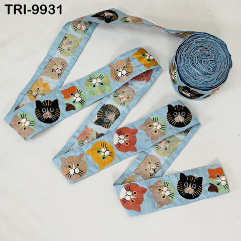 a blue fabric with a repeating pattern of cat faces in various colors, including black, orange, and green. The fabric is rolled up and placed on a white surface, with a spool of the same blue fabric visible in the background.