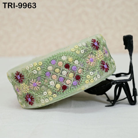 a decorative green fabric with a floral pattern, including purple and pink flowers, arranged in a circular design. The fabric is displayed on a white surface, and there is a small black metal sculpture or figurine positioned next to it.